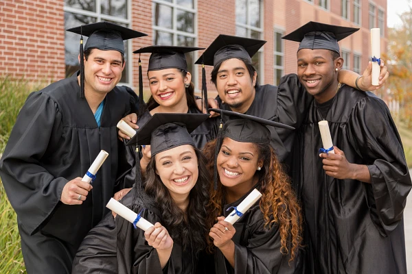 6 young adults wearing graduations caps and gowns, holding rolled-up diplomas, smiling