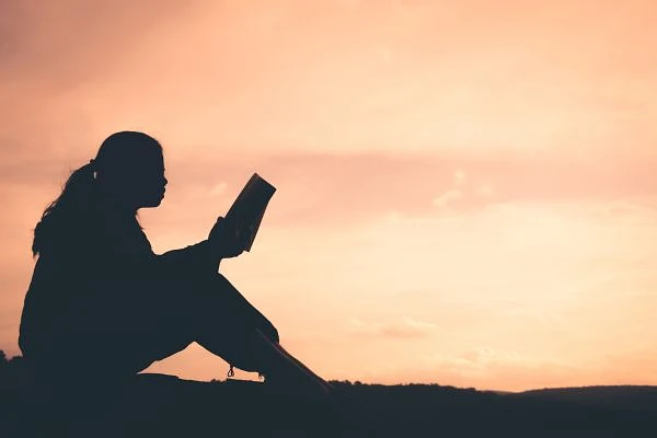 A person is sitting outdoors, silhouetted against a sunset sky, reading a book.